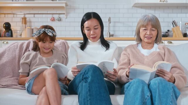 Asian grandmother, mother, and little girl are sitting together on a sofa, each reading their own book