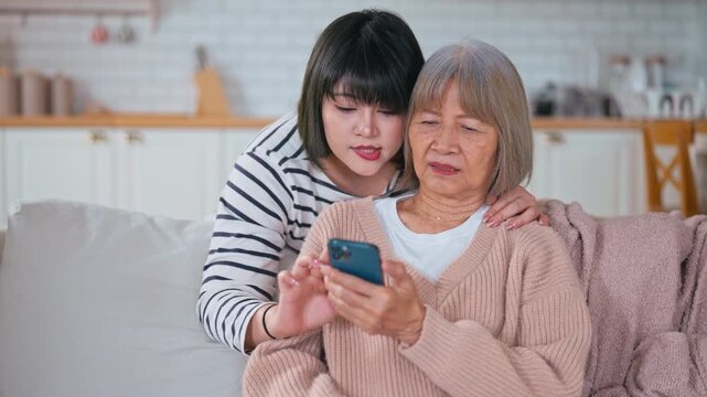 Asian mother and her daughter sit on a sofa, looking at a smartphone together. The daughter teaches her mother how to use social media, shop online, and view family pictures in a cozy home