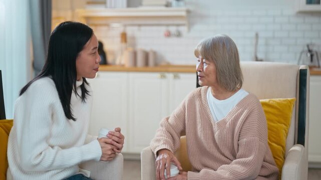 Asian adult daughter listens attentively while holding hands with her elderly mother over coffee. They are sharing a moment of emotional support in a cozy, warm living room at home together.