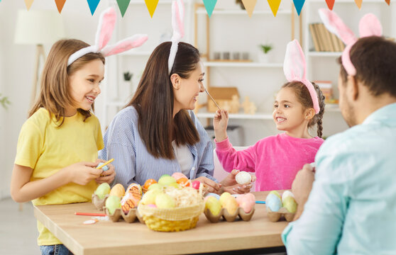 Family portrait of happy mother and father with little cute children in bunny ears sitting at table in kitchen with painted multi-colored Easter eggs. Holiday preparation and celebration concept.