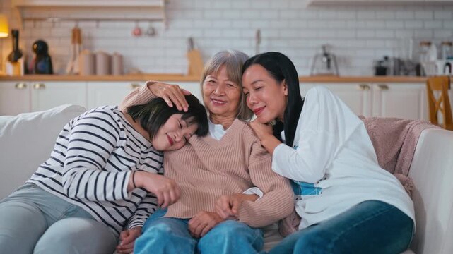 Happy Asian mother, sister, and daughter hugging closely on a sofa, showing love, family bonding, and warmth in a cozy home environment.