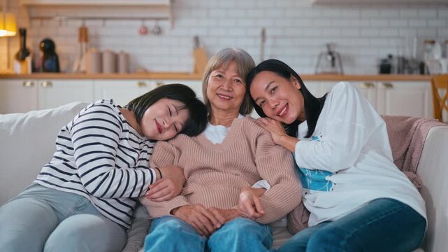 Slow motion Happy Asian mother, sister, and daughter hugging closely on a sofa, showing love, family bonding, and warmth in a cozy home environment.