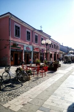 Street view of historic pink building with outdoor cafe seating red chairs bicycle parked on cobblestone pavement and clear blue sky during a sunny day afternoon. Shkoder, Albania - September 23, 2017