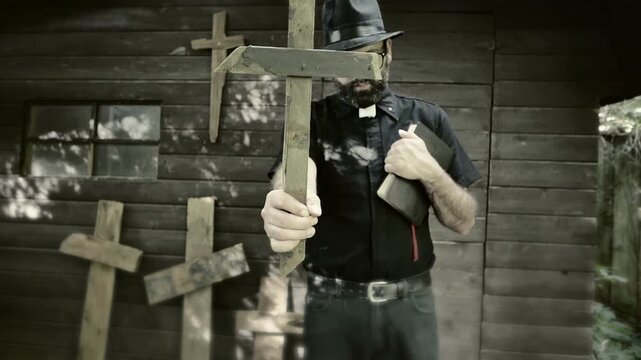 Clergyman, reverend or priest wearing a clerical collar, holding a bible and a wooden crucifix. Preacher preaching the gospel in front of an old rustic rural church.