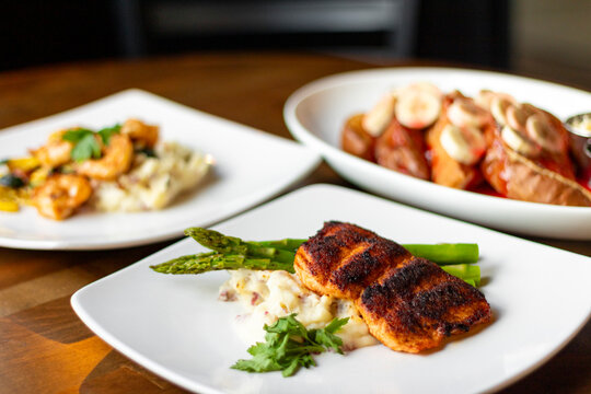 Three white plates on a wooden table featuring an assortment of foods from shrimp and salmon to french toast and bananas, as available in a diner or restaurant.