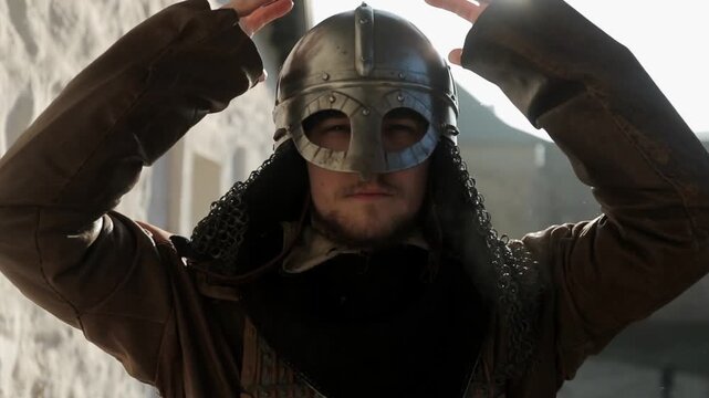 Dramatic steadicam push-in shot of Viking warrior putting on metal helmet, camera moving from mid shot to close-up in sunlit stone fortress. Cinematic battle preparation historical reenactment footage