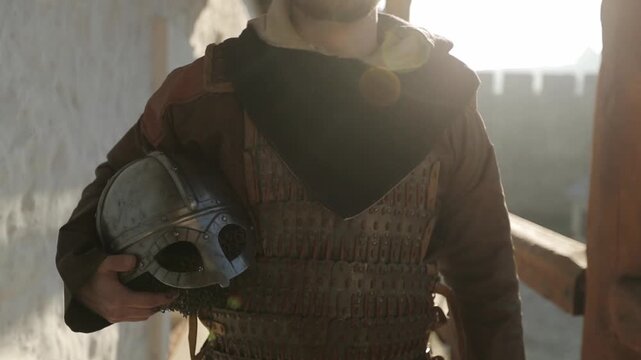 Bearded Viking warrior in chainmail armor holding helmet walking confidently toward camera through sunlit medieval stone fortress. Dramatic cinematic historical reenactment footage for history and cul