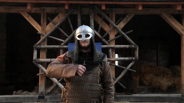 Bearded man in Viking helmet and chainmail armor walking toward camera making cross sign, with Ukrainian flag visible in background inside wooden medieval hall. Historical patriotic cultural content
