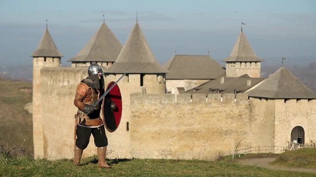 Full body wide shot of Viking warrior in armor holding sword and red shield standing before impressive medieval castle with multiple towers under blue sky. Epic historical reenactment location footage