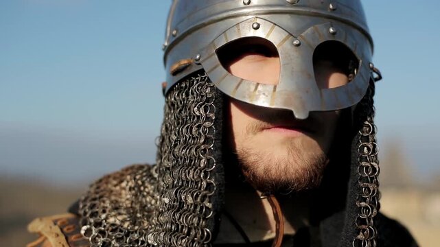 Close-up portrait of bearded Viking warrior in polished metal helmet and chainmail coif against bright blue sky. Powerful cinematic historical reenactment character shot for history and fantasy conten