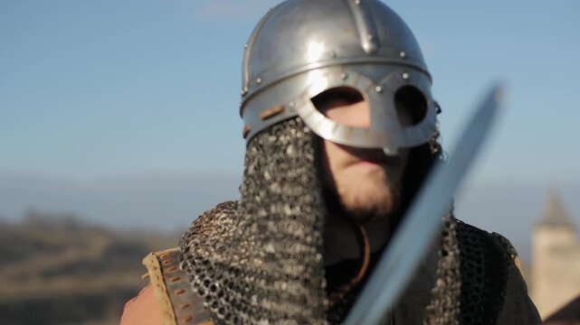 Viking warrior in metal helmet and chainmail striking sword against shield and gazing into the distance against blue sky. Dramatic battle-ready cinematic shot for historical reenactment content