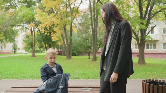 Mother gets ready. Child and mother prepare together. Morning ritual of mother and son in autumn setting. Woman helps young boy dress in park amidst falling leaves before school starts