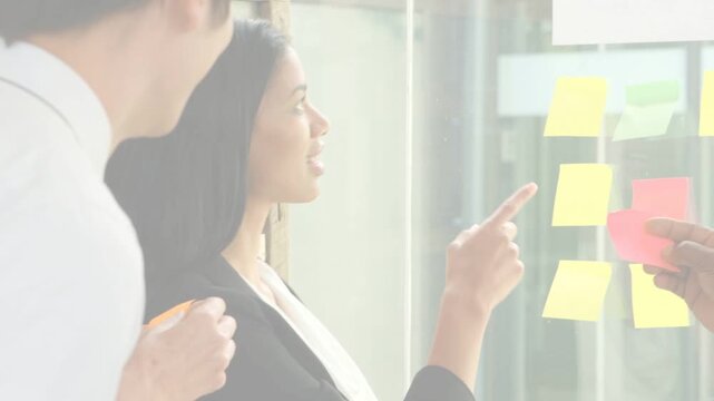 Business lead pointing at glass wall while colleague handing pink sticky note, prioritizing ideas