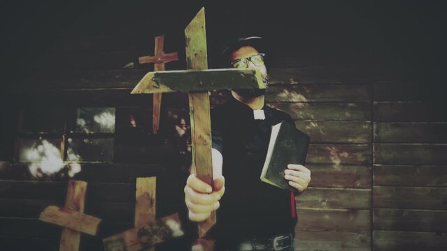 Clergyman, reverend or priest wearing a clerical collar, holding a bible and a wooden crucifix as if conducting an exorcism. Preacher preaching the gospel in front of an old rustic rural church.