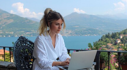 Obraz premium Focused young woman in white shirt works on her laptop on balcony overlooking beautiful lake and mountains. High quality photo
