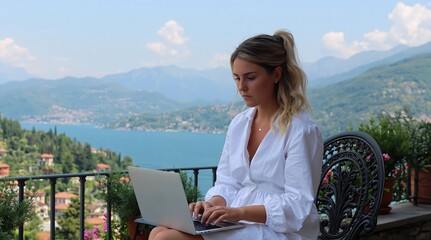 Obraz premium Woman in white dress works on her laptop on balcony overlooking beautiful lake and mountains. High quality photo