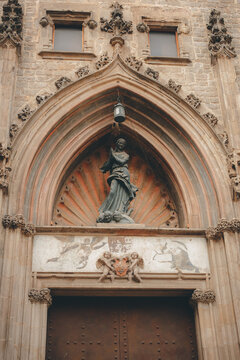 Gothic facade detail of an ancient church in Barcelona, Spain. Stone portal with a religious statue of the Virgin Mary, ornate carvings, and historic wooden doors in the Gothic Quarter (Barri Gotic).