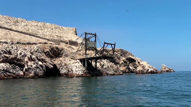 Cinematic boat pass-by of coastal pier structure and rocky shoreline with seabirds near Ballestas Islands, Paracas National Reserve, Peru