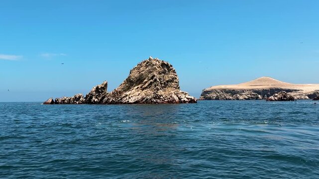 Cinematic wide establishing shot of rocky islet formations with foreground sea rock and distant island cliffs, Isla Centro Chimcha near Paracas National Reserve, Peru, with flying seabirds