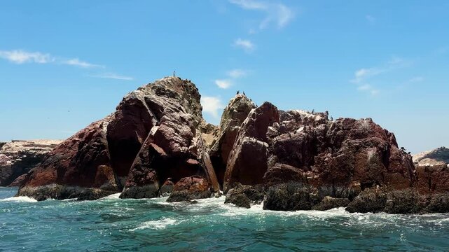 Cinematic boat pass-by of rugged rocky sea stack with waves breaking and seabirds perched, coastal island formations near Paracas National Reserve, Peru