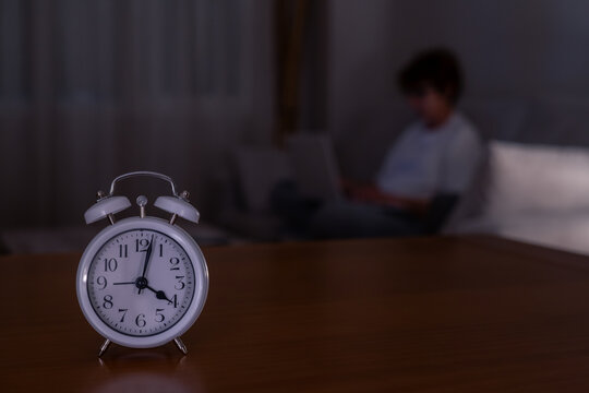 Alarm clock reads 4:00 am on a table in a dark room, blurry laptop and lone worker in the background conveying sleeplessness, late-night work, exhaustion and burnout