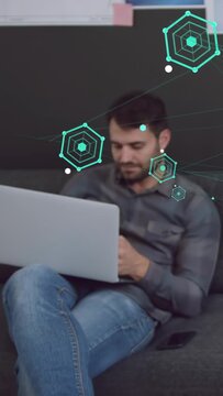 Vertical video: IT pro tapping phone, causing teal hexes over face and laptop while reviewing data