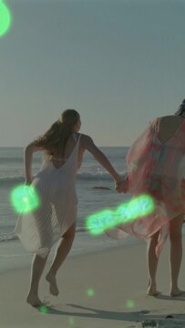 Vertical video: Two women raising hands skipping along beach in joy, green orbs boosting travel ad