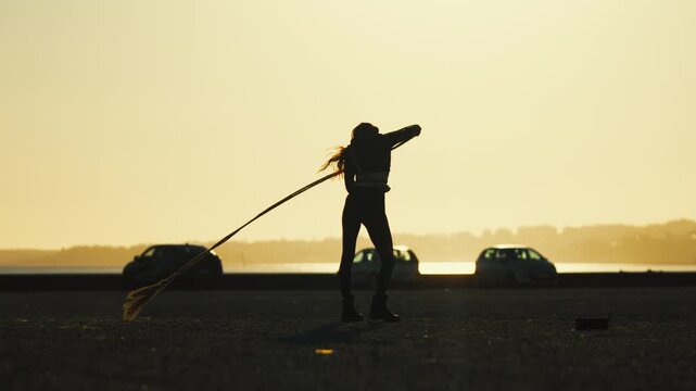 Woman practices hula hoop dance by the water during sunset with cars parked nearby