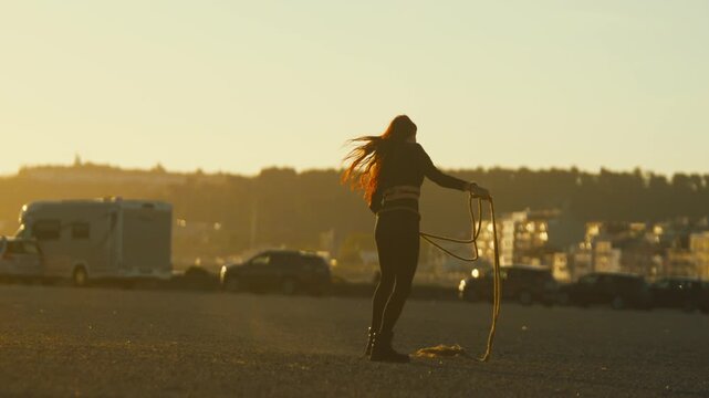 Woman practices lasso skills at sunset by the beach with cars in the background