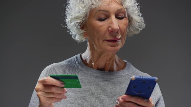 Old woman holds smartphone and card in hands. Grandmother buys groceries online. Senior female enters bank details into delivery application. Retired lady makes purchases, buys tickets