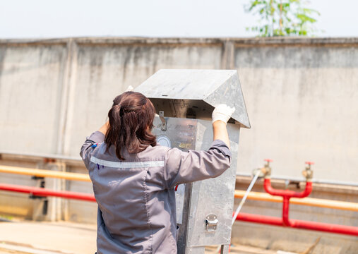 A technician in a gray industrial uniform inspecting an outdoor environmental monitoring station or air quality sampling box at a treatment facility.