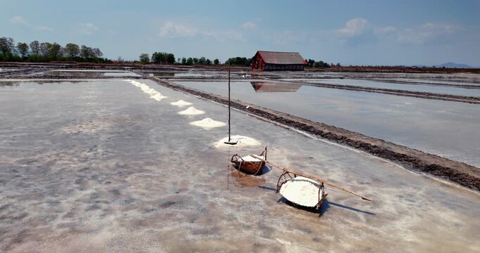 Panning shot of traditional salt pans featuring woven baskets on a carrying pole, harvested salt mounds, and a wooden storage shed reflecting in the water.