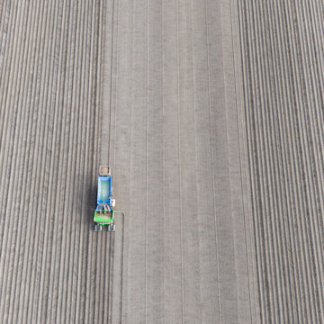 Aerial view of a blue and green agricultural machine planting seeds in a large field with straight vertical furrows.