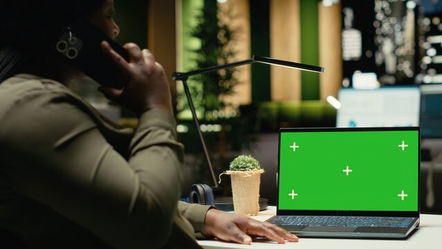 Isolated green screen on laptop next to black woman answering a phone call, handling administrative tasks after hours at night. Business manager works on agenda development. Camera B.
