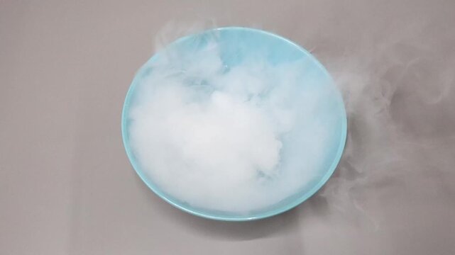 Top view of liquid nitrogen dry ice smoke bubbling in blue bowl on neutral background.