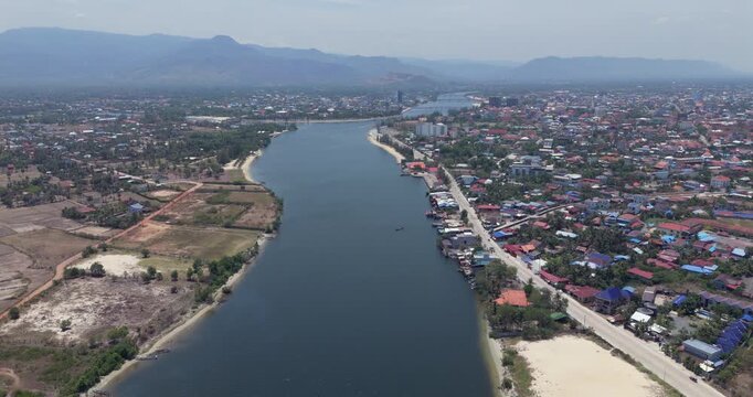 Forward aerial view of the Kampot river flowing alongside a town with colorful rooftops, beneath distant hazy mountains.