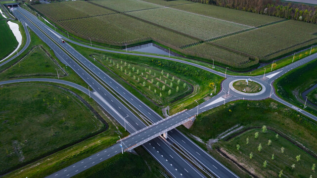 Aerial view of a complex highway interchange featuring a roundabout and a bridge over multiple lanes of traffic adjacent to lush green orchards and agricultural fields.