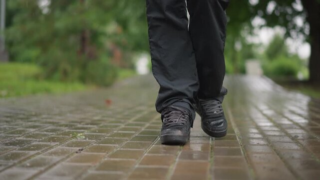Wet brick path woman walking slowly, closeup low angle of black boots stepping through shallow puddles on glossy pavement overcast park in background with trees and damp greenery, calm contemplative
