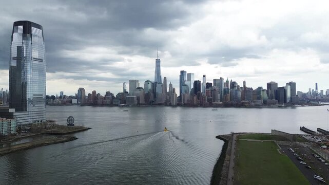 Panoramic skyline sweeps captures Hudson River corridor between Liberty State Park and Lower Manhattan with One World Trade Center anchoring dense urban backdrop. Lawn textures, walkway lines, NJ
