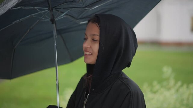 Joyful rainy outdoor. Candid woman under rain umbrella smiling happily. Female enjoying cheerful moment in rainy park setting. Woman with hooded coat smiling joyfully as rain gently falls in park