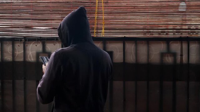 Man in black hoodie behind curtain using phone to type messages or send secret email. Mysterious person indoors holding smartphone. Hacker or spy in a house.