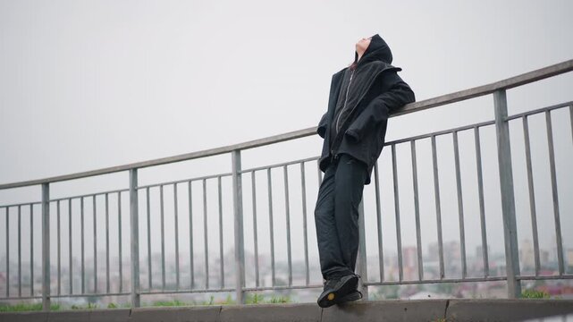 Rainy city release. Drenched woman relaxes against railing outdoors. Cityscape woman breathes freely in rainy urban scene. Wet pavement and distant skyline frame woman finding relief in rain