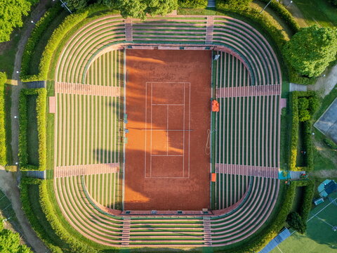 Aerial view of the red clay tennis court at Circolo della Stampa Sporting surrounded by tiered grass seating and lush green hedges in Torino, Piemonte, Italy.