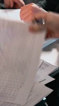 A focused closeup shows a hand writing on a document at a desk, with a pen over a grid sheet, conveying formal paperwork, contract, and business activity in an office setting