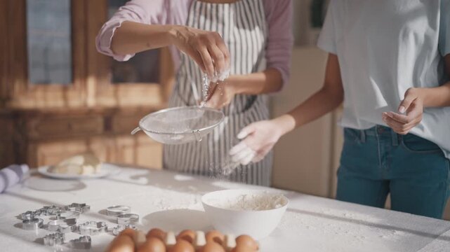 Middle-aged and young women cooking together in home kitchen, sifting flour. Calm family weekend, mother and daughter making dough for cookie, pie, pizza or pasta for fest dinner, details shot