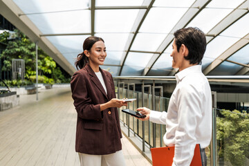 Obraz premium Two professionals in a modern atrium use a smartphone and access card for a contactless check-in, smiling during a casual workplace interaction surrounded by glass and greenery