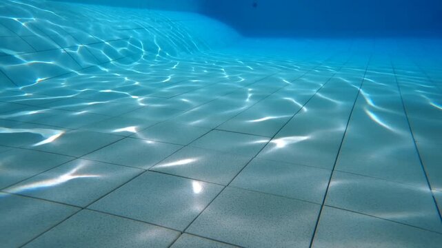 Slow motion static pan of tiled pool floor with shifting light caustics underwater in indoor swimming facility