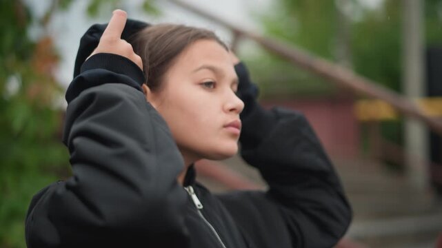Side profile woman pulling hoodie up, damp stairway and metal railing in background, thoughtful expression while adjusting zipper and hood, anticipatory mood with urban steps and subtle drizzle roles
