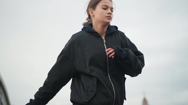 Young woman crossing rainy market street, steady stride through parked cars, hoodie zipped, urban storefronts and cloudy sky, candid walking motion, purposeful expression, editorial energy