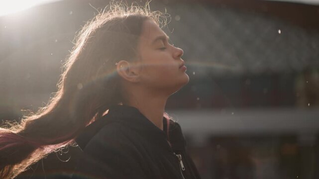 Young teen girl backlit urban portrait, soft golden rim light, loose braid, dark hoodie, pensive gaze toward sky, soft bokeh background, light flare and dust motes, subtle wind through hair, cinematic